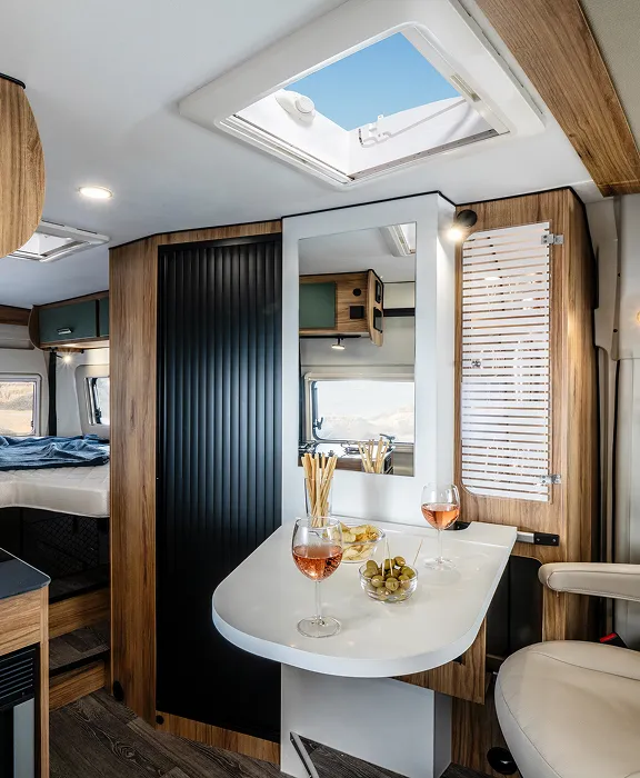 Moderner Wohnmobil-Innenraum mit Holzelementen, weißem Tisch, gedrehtem Beifahrersitz und Blick auf das Heckbett und Meer.